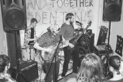 Black and white photo of three musicians playing drums, guitar, and bass in a room with two big speakers in front of a sign that reads “-AND TOGETHER DENTON.” The backs of several audience members’ heads can be seen.