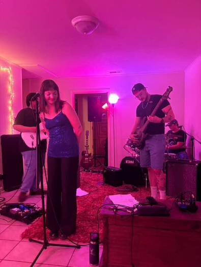 A drummer, bassist, and guitarist stand behind a singer in a room with purple lighting.