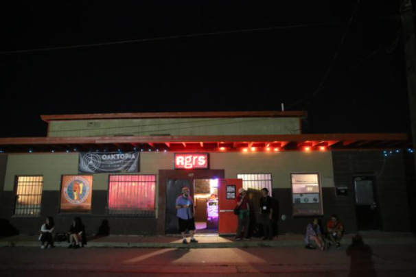 A building against a night sky with a red neon sign that says “rgrs.” Several people stand outside or sit on the curb in front of the building.