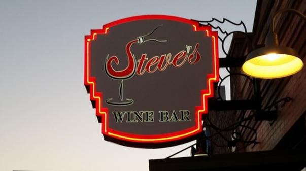 A sign attached to a building. The sign says “Steve’s Wine Bar” and the “S” in Steve is designed to look like wine flowing from a bottle into a glass.
