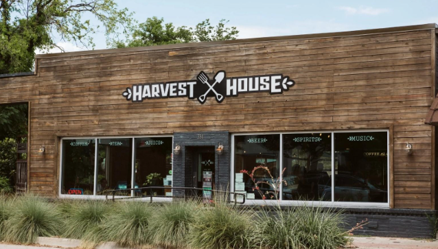 A building with a sign that says “HARVEST HOUSE” with a logo of a shovel and a garden fork tool between the words. There are bushes in front of the building and it is daytime.