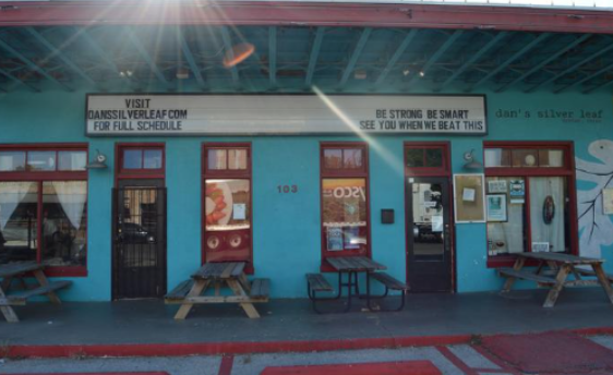 The front of a building that is painted light blue with four picnic tables out front and a sign above the doors that say “Visit danssilverleaf.com for full schedule” and “Be Strong Be Smart See You When We Beat This.”