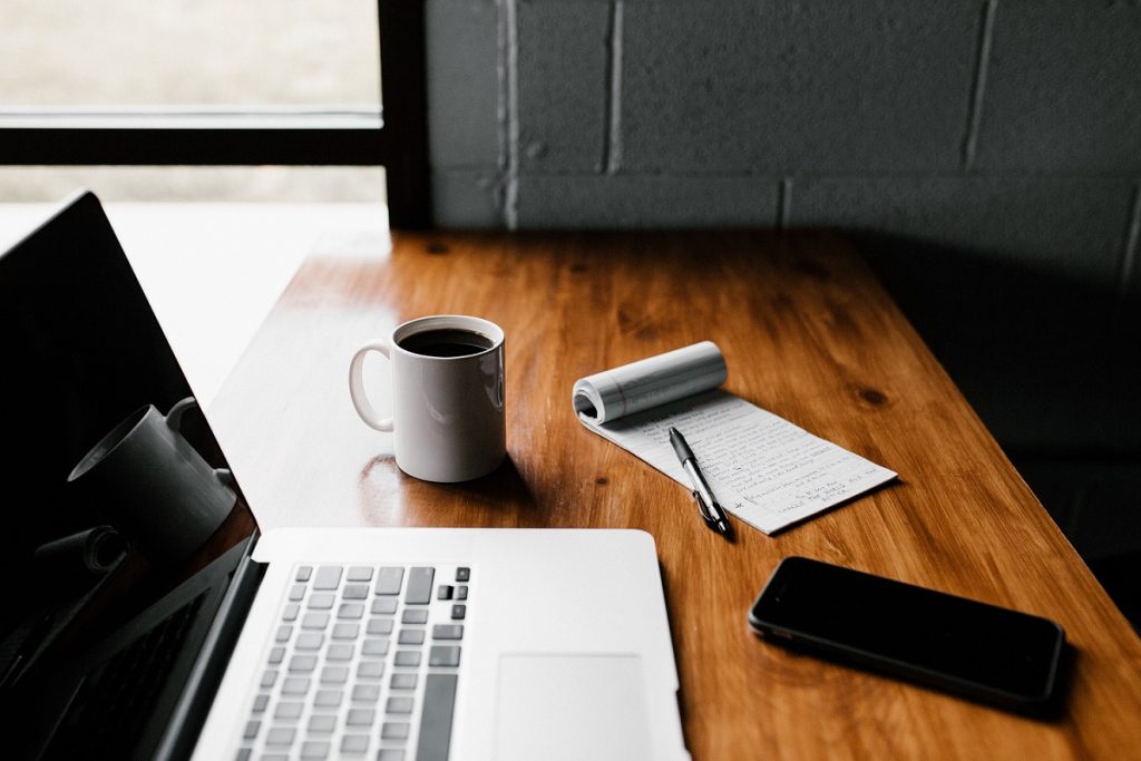 Laptop on a table next to a coffee, phone and notepad