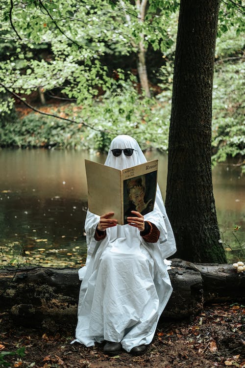 A person wearing a white sheet and sunglasses, viewing an ancient-looking book while sitting under a tree in a forest. 