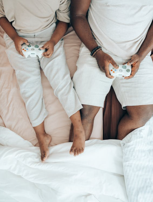 Two people laying in bed and playing a video game together. Both of them are holding video game controllers in their hands. Their heads cannot be seen. 