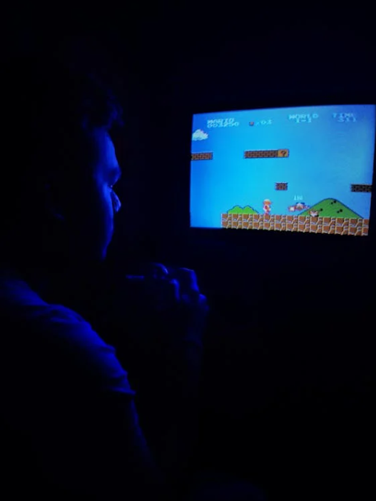A person in a dark room is playing the video game Super Mario Brothers on a screen. The person’s features are illuminated in a dark blue light. 