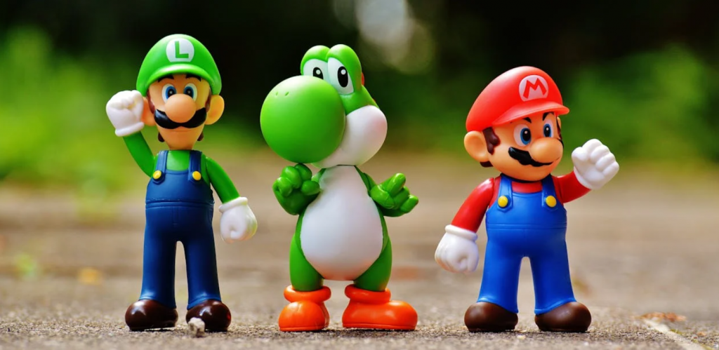 Toy figures of (L to R) Luigi, Yoshi, and Mario stand side by side on a gravel road. 