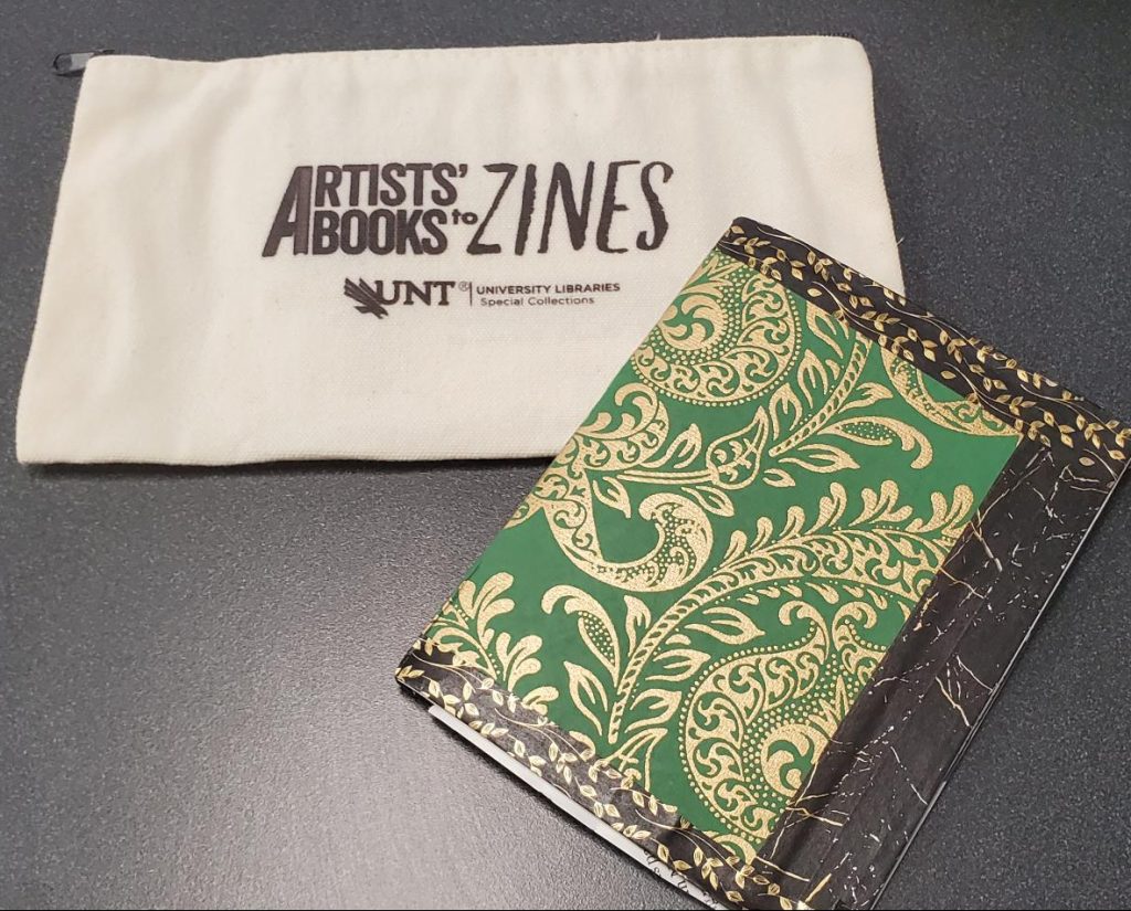 A green and gold patterned booklet with black and gold bordering in front of a pen pouch that says "Artists' Books to Zines'.
