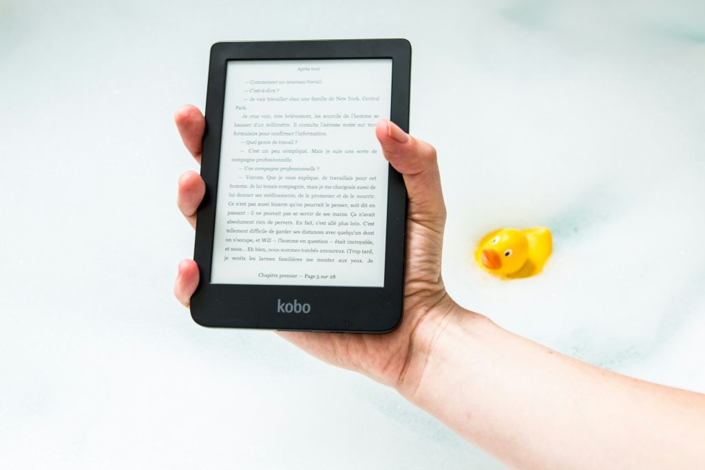 Outstretched hand holding an e-reader over a bubble bath with a rubber ducky.