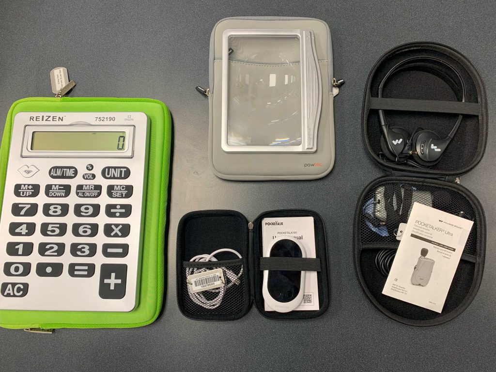 Large green calculator, rectangular plastic magnifier, headphones in a case, and a small hand-sized white device.