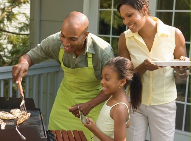 An African American father, mother, and young daughter barbecuing in the backyard.