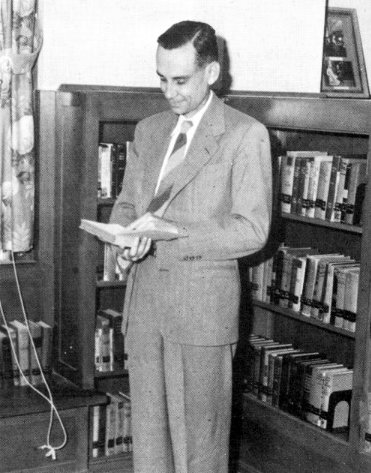 Photo from the 1948 edition of The Yucca, Yearbook of North Texas State College. Library Director Arthur M. Sampley is looking down at a book in his hands while standing in front of a shelf full of books behind him.
