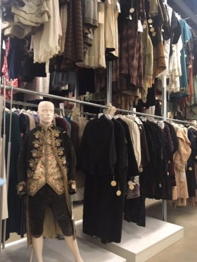 Color photograph of a mannequin in old fashioned clothes standing at the end of a two-tiered hanging rack full of clothes.