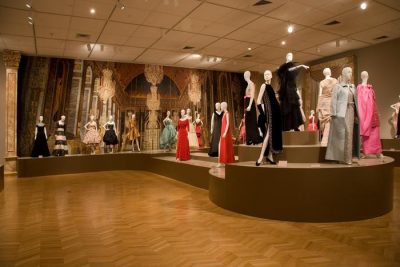 Color photograph of a large room with a riser along the back wall and coming into the center of the floor. Mannequins in evening gowns are displayed along the riser.