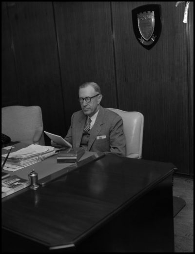 Black and white photograph of older white man sitting at a desk earing a suit.