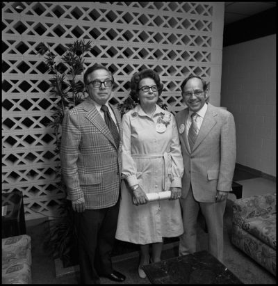 Black and white photograph of a woman standing between two men posing for a photo. The men wear suits, while she wears a white dress.