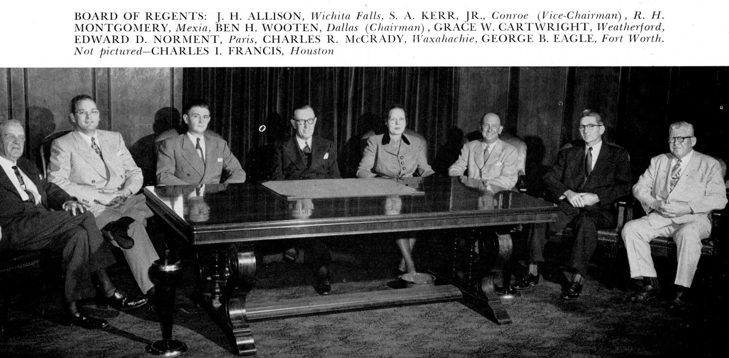 Photograph of eight people sitting behind a large wooden table wearing suits. All are white men except for one white woman.